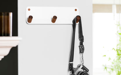 The Plane 3 Wall Hook by Fire Road, a white wall-mounted rack made of sturdy powder-coated steel with three wooden pegs, securely holds a camera strap with a camera attached.