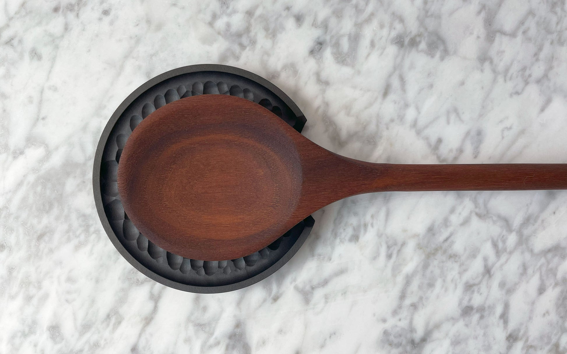 A sustainable Chisel Spoon Rest by Fire Road rests on a black silicone trivet placed on a gray marble surface.