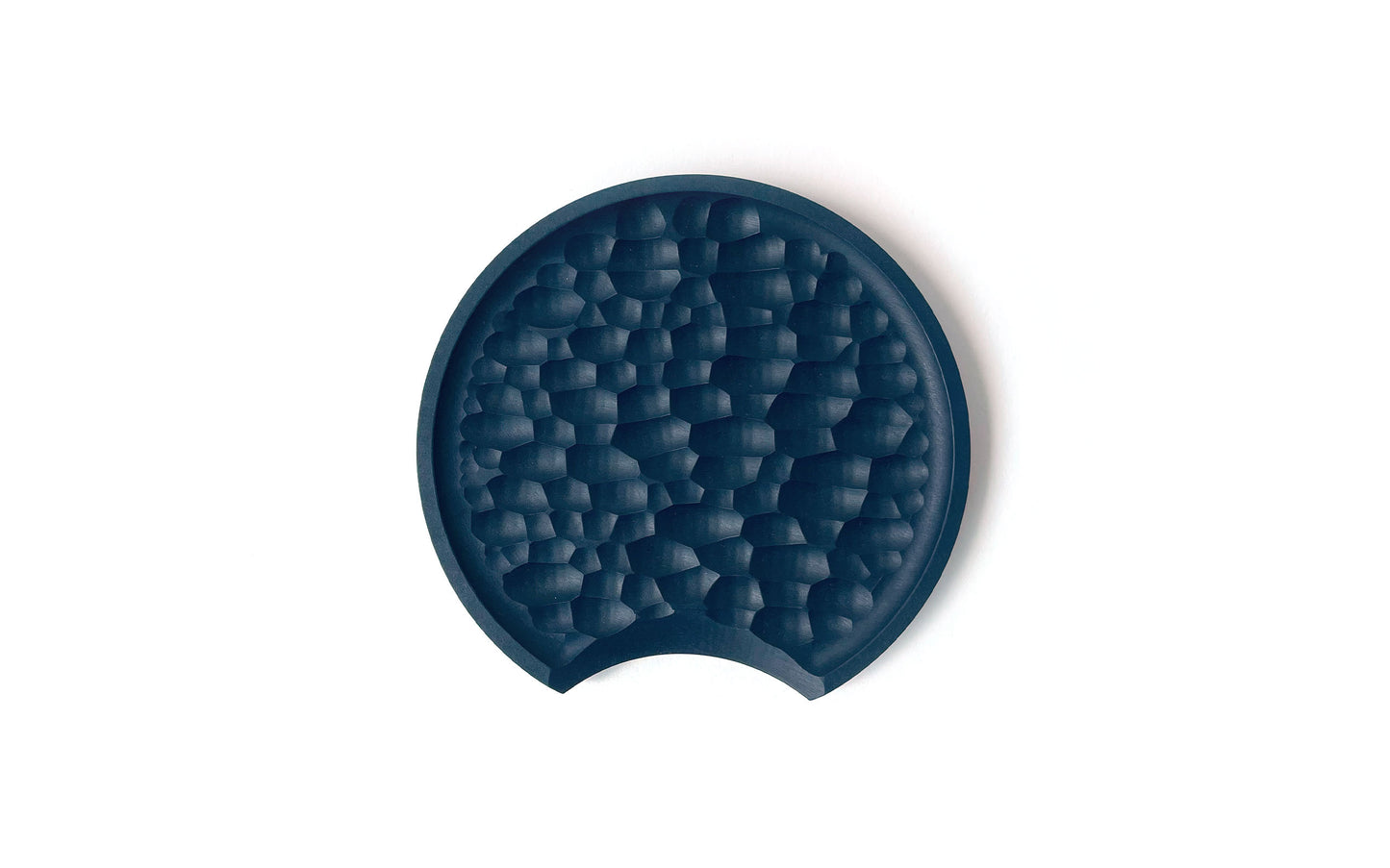 A round, dark-colored tray made from recycled paper kitchenware features a textured, geometric pattern inside and a small notch on one edge. It’s placed against a white background. This is the Chisel Spoon Rest by Fire Road.