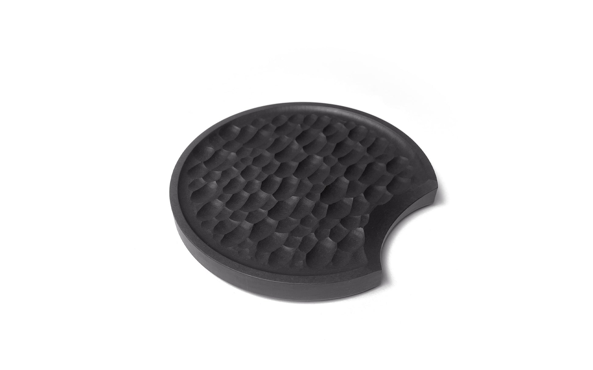 A round, black textured kitchen utensil with a small crescent-shaped notch on one side, crafted from sustainable recycled paper, shown against a plain white background— the Chisel Spoon Rest by Fire Road.