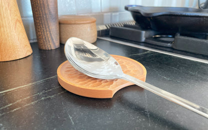 A stainless steel spoon rests on a Chisel Spoon Rest by Fire Road, placed on a textured wooden holder on a black kitchen countertop next to the stove.